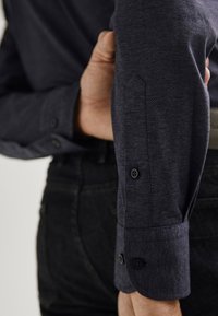 Camicia blu navy con tessuto testurizzato. Presenta una vista ravvicinata della manica con polsini a doppio bottone e un design aderente.