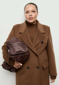 Bruine wollemix overjas met een dubbelgeknoopte ontwerp, gecombineerd met een gestructureerde bruine gebreide trui, terwijl hij een donkerbruine leren rugzak vasthoudt.