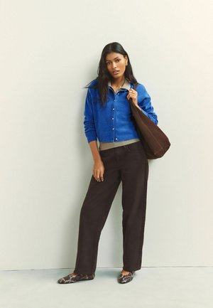 Naine, kes kannab sinist kardigani, halli särki, tumedaid laia säärega pükse ja käib madalatel kingadel, millel on madu-mustriga muster, hoides suurt pruuni poekotti õlal.