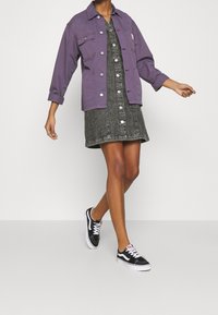 Purple cotton jacket with front pockets layered over a dark denim dress featuring button closure. Paired with black and white sneakers.