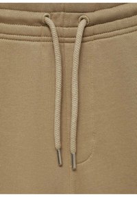 Pantalons de survêtement beige avec une taille élastique et un cordon de serrage. Texture douce, avec des embouts en métal et une poche latérale.
