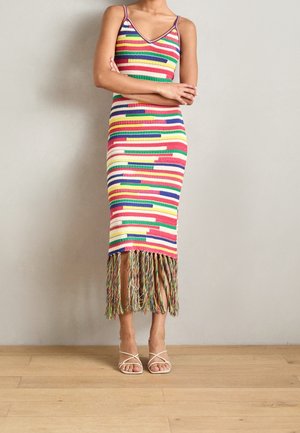 Vrouw staat met gekruiste armen en draagt een kleurrijke, gestreepte gebreide jurk met spaghettibandjes en franjes aan de zoom, gecombineerd met witte sandalen met bandjes.