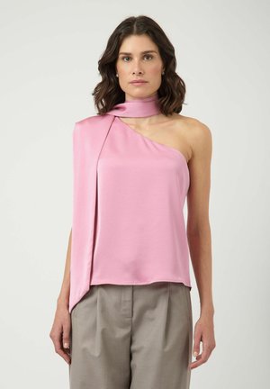 Femme portant un top en satin rose sans manches avec une épaule découverte et un pantalon taille haute couleur taupe, debout devant un fond uni.