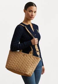Tan quilted tote bag with chevron pattern, dual handles, and adjustable shoulder strap. Features a subtle logo on the front.
