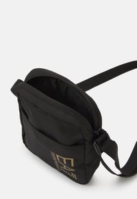 Borsa a tracolla in tessuto nero con compartimento principale con zip, tasca frontale piatta, tracolla regolabile e dettagli del logo dorati sulla parte anteriore.