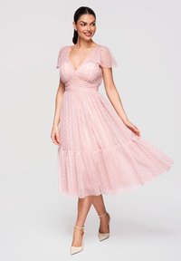 Femme souriante, portant une robe rose pâle longueur genou à pois, manches papillon, et des talons blancs avec bride à la cheville, debout devant un fond uni.