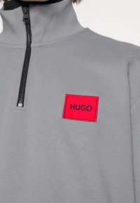 Šedý pulovr na zip s vysokým límcem, vybavený černým zipem. Červený obdélníkový nášivka s textem "HUGO" na levé straně. Hladký povrch.