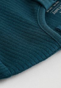 Tessuto a coste color teal con una consistenza morbida, caratterizzato da un colletto rotondo e un orlo curvo. Cuciture dettagliate sono visibili lungo i bordi.