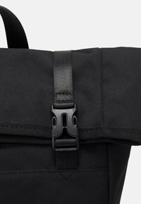Sac à dos en tissu noir avec une boucle en plastique à déclenchement latéral sécurisant une fermeture à rabat supérieur, vu de près.