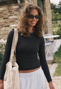 Zwart geribd longsleeve-top, lichtgrijze joggingbroek en een beige schoudertas met een band waarop het woord "SELF" staat. Krullend haar en zonnebril.