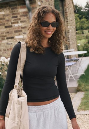 Top nero a maniche lunghe a coste, pantaloni della tuta grigio chiaro e una borsa a tracolla beige con una cinghia con la scritta "SELF". Capelli ricci e occhiali da sole.