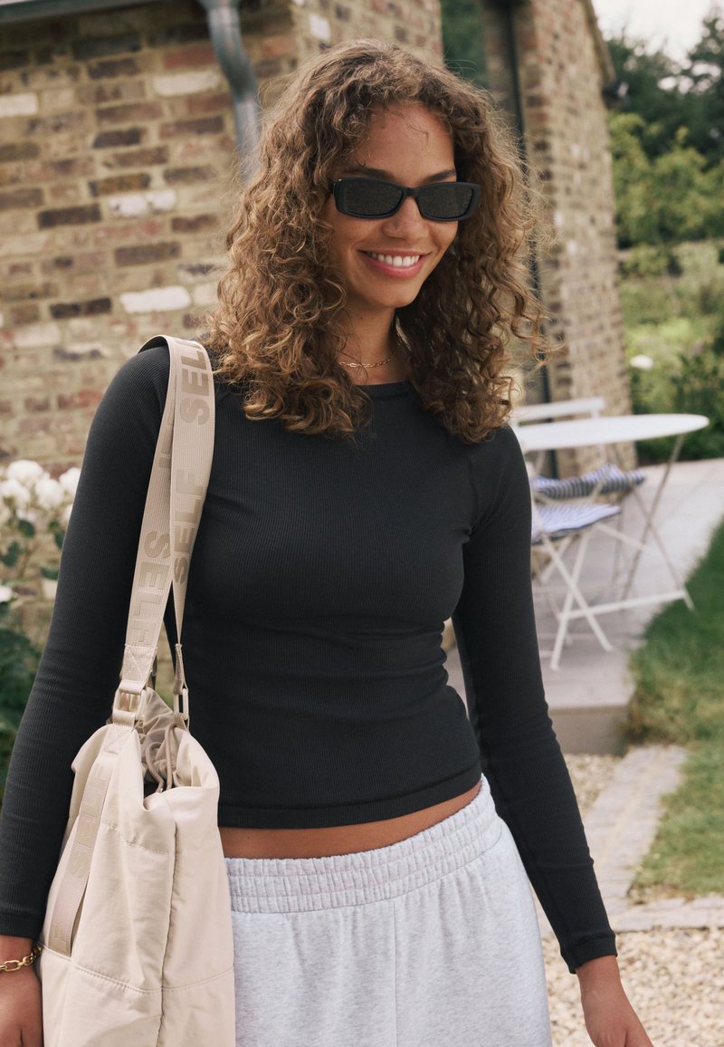 Zwart geribd longsleeve-top, lichtgrijze joggingbroek en een beige schoudertas met een band waarop het woord "SELF" staat. Krullend haar en zonnebril.
