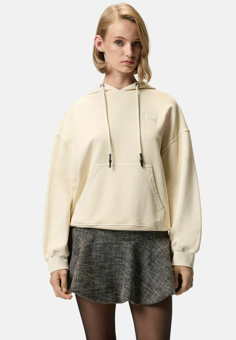 Felpa color crema con tasca a canguro, cappuccio con cordino e logo ricamato. Indossata sopra una gonna grigia a trama.