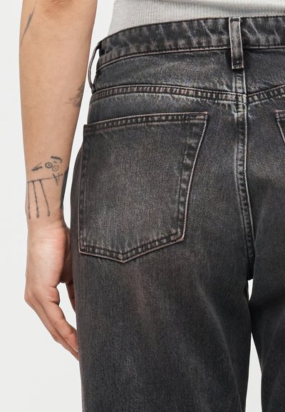 Αριστερό χέρι με μαύρη γραμμή και αφηρημένα τατουάζ δίπλα στο πίσω μέρος ατόμου που φοράει ξεθωριασμένο μαύρο τζιν και γκρι πουκάμισο.