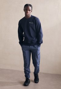 Sweat-shirt bleu marine avec un logo sur la poitrine, associé à un pantalon cargo bleu foncé avec des zippers, et des chaussures noires. Fond à surface plane.