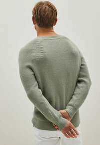 Grijs-groene gebreide trui met een ribbelstructuur, lange mouwen en een relaxte pasvorm, gezien van achteren met de handen achter de rug geplaatst.