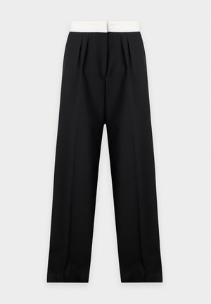 Femme aux longs cheveux foncés portant un haut gris à manches longues, un sous-vêtement blanc, un pantalon large noir et des chaussures noires, debout sur un fond blanc.