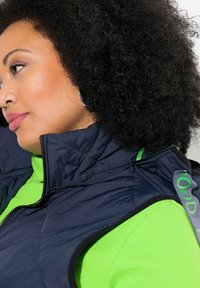 Granatowa kamizelka puchowa z wysokim kołnierzem, z kieszeniami na zamek i zielonymi akcentami, noszona na jaskrawo zielonym sweterku z prążkami.