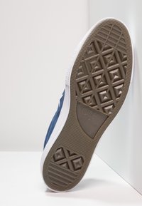 Zapatilla de lona azul con suela de goma blanca, que presenta una suela exterior marrón texturizada y un patrón triangular para tracción. Conlleva la marca "Converse All Star".