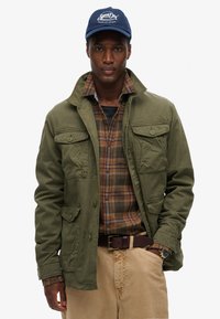 Grøn bomuldsjakke med fire forlommer, ternet skjorte under, beige bukser og en blå kasket med logo-tekst.