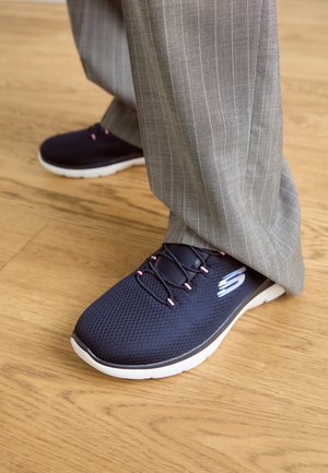 Granatowe sneakersy Skechers z różowymi akcentami noszone z szarymi spodniami w cienkie prążki na drewnianej podłodze.