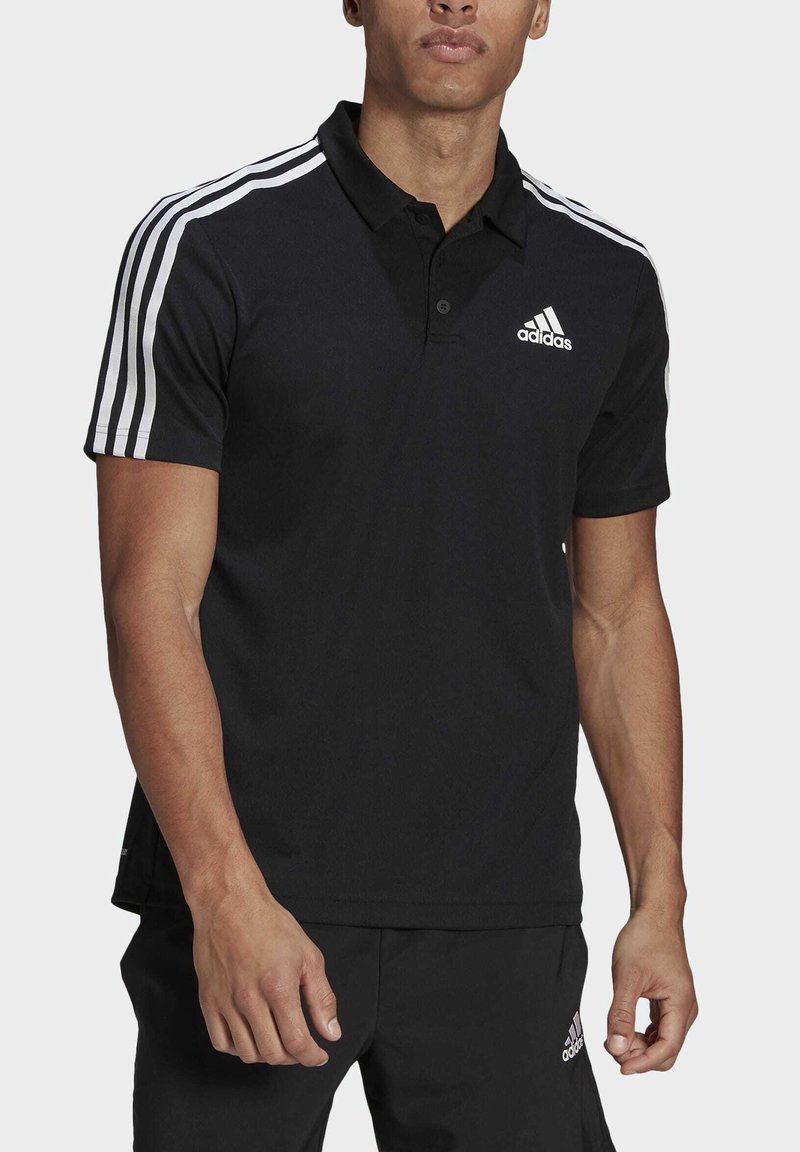 Homme portant un polo noir Adidas avec des bandes blanches sur les manches et un pantalon Adidas noir assorti, debout devant un fond uni.