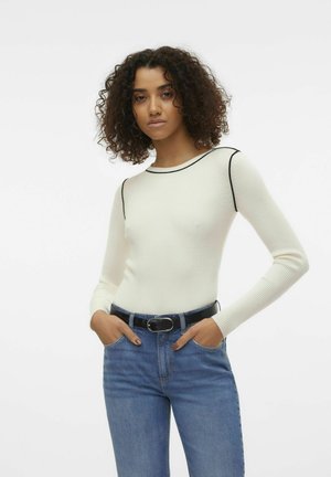 Maglione bianco a coste con maniche lunghe e rifiniture nere sul colletto, abbinato a jeans blu e una cintura nera con fibbia argentata.