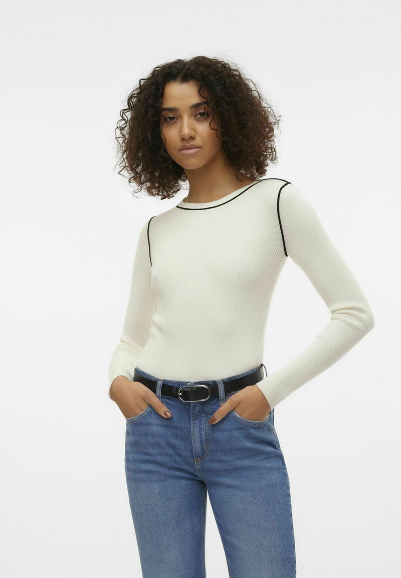 Maglione bianco a coste con maniche lunghe e rifiniture nere sul colletto, abbinato a jeans blu e una cintura nera con fibbia argentata.