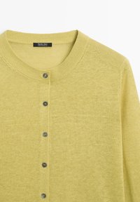 Cardigan jaune pâle boutonné avec col ras du cou et manches longues, présentant une petite étiquette noire à l'intérieur du col.