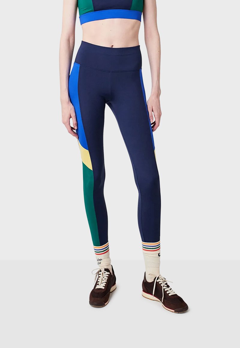 Legíny s barevnými bloky v námořnické modré, modré, zelené a žluté barvě, nošené s bílými pruhovanými ponožkami a tmavě hnědými šněrovacími teniskami.