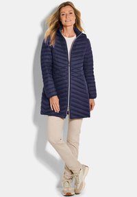 Marineblauwe gewatteerde jas met een hoge kraag en volledige frontale rits, gecombineerd met lichtbeige broek en crèmekleurige sneakers. Soepele textuur, lichtgewicht ontwerp.