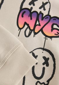 Gros plan sur un tissu beige avec un bord-côte, des lettres style graffiti en violet et orange, et le contour noir d'un visage souriant avec des yeux barrés.