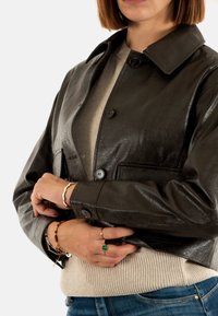 Giubbotto in pelle marrone scuro con colletto, bottoni frontali e due tasche laterali, abbinato a un maglione beige a coste.