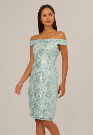 Kobieta ubrana w bladoniebieską sukienkę odsłaniającą ramiona, sięgającą do kolan, zdobioną błyszczącymi wzorami kwiatów i winorośli, stojąca na jednolitym tle.