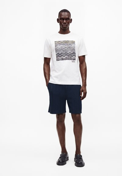 Homme portant un t-shirt blanc avec un motif carré ondulé et du texte, un short bleu marine texturé et des mocassins noirs, debout sur un fond blanc.