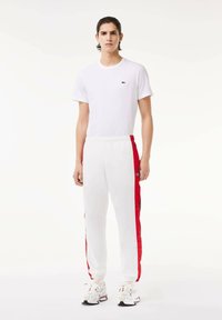 Balta marškinėliai su mažu žaliu logotipu, balti sportiniai džinsai su raudonais šonais ir balti sportbačiai su juodu akcentu. Lygus audinys ir laisvas siluetas.