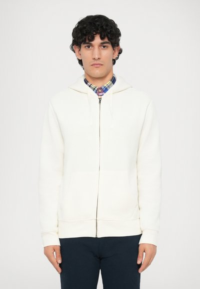 Man met krullend donker haar die een witte zip-up hoodie over een meerkleurig geruite shirt en donkere broek draagt, staand tegen een effen achtergrond.