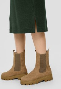 Brązowe zamszowe botki za kostkę z siateczkowymi panelami bocznymi i masywnymi gumowymi podeszwami. W połączeniu z ciemnozieloną sukienką do kolan z rozcięciami po bokach.