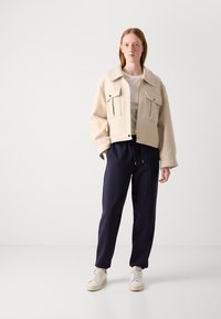 Mladá osoba stojí, má na sobě béžovou bundu s velkými kapsami, bílou košili, tmavě modré kalhoty se stahovací šňůrkou a bílé tenisky na bílém pozadí.