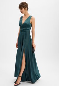 Groene satijnen avondjurk met een diepe V-hals, aansluitende taille en hoge split; gecombineerd met een zwarte clutch en sandalen met hoge hak en bandjes.