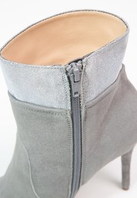 Szary zamszowy botek za kostkę z srebrną tkaninową kołnierzem, bocznym zamkiem błyskawicznym i szpilką. Gładka tekstura z dopasowaniem wokół kostki.