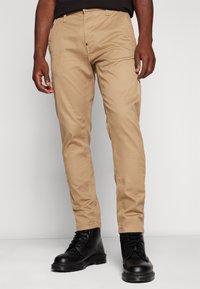 Beige chinos iz bombaža, s slim fit krojem in petimi žepi. V kombinaciji s črnimi čipkastimi škornji za kontrast.