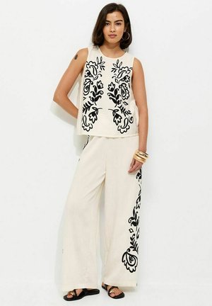 Mujer con blusa blanca sin mangas y pantalones de pierna ancha con motivos florales negros, sandalias negras y brazaletes dorados en una muñeca.