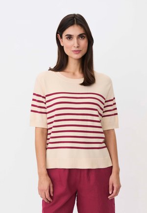 Kobieta o prostych ciemnych włosach ubrana w beżowy sweter z krótkim rękawem z poziomymi czerwonymi paskami i czerwone spodnie, stojąca na tle jednolitego białego tła.