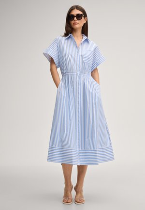 Vrouw die een midi-jurk draagt met blauwe en witte verticale strepen, korte mouwen, een kraag, een borstzakje aan de voorkant en een elastische taille, gecombineerd met een zonnebril.