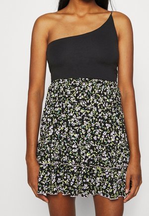 Frau trägt ein schwarzes Ein-Schulter-Oberteil kombiniert mit einem kurzen schwarzen Rock mit einem kleinen weißen und grünen Blumenmuster.