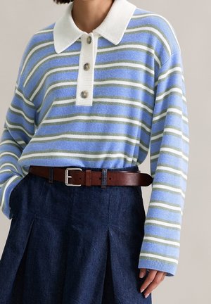 Frau trägt einen blau-weiß-grau gestreiften Langarm-Polo-Pullover, in einen dunkelblauen Jeansrock mit braunem Gürtel gesteckt, eine Hand in der Tasche.