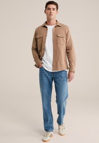 Rudas mygtukų marškinis su dviem krūtinės kišenėmis, uždedamas ant baltos marškinėlių, derinamas su šviesiai mėlynomis džinsomis ir kreminiais sportbačiais su rudais akcentais.