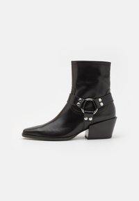 Bottine en cuir noir avec un bout pointu, un talon moyen et un strap décoratif avec un accent métallique circulaire. Texture lisse.