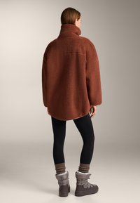 Brown fleece oversized jacket with high collar, textured surface, and rounded hem. Worn over black leggings and gray winter boots.